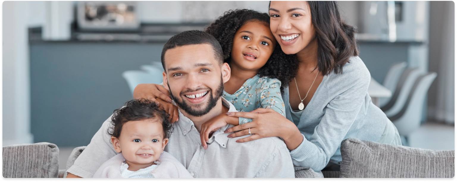 A family of four, with a mother, father, toddler, and baby, smiling and posing together on a couch in a living room.